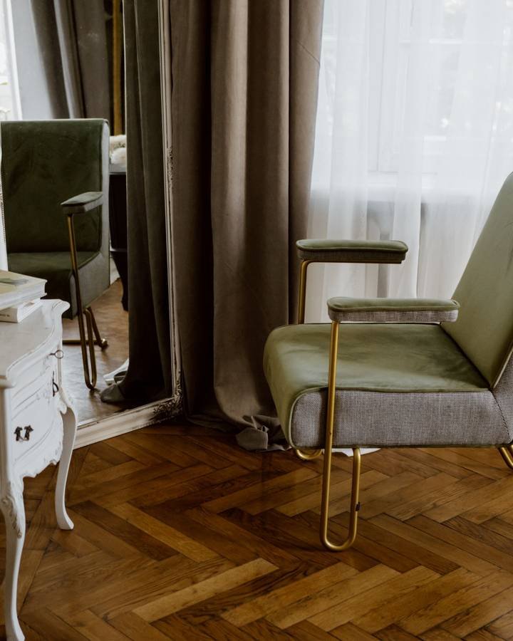 A sage velvet reading chair with brass legs beside floor-length drapes on a herringbone floor