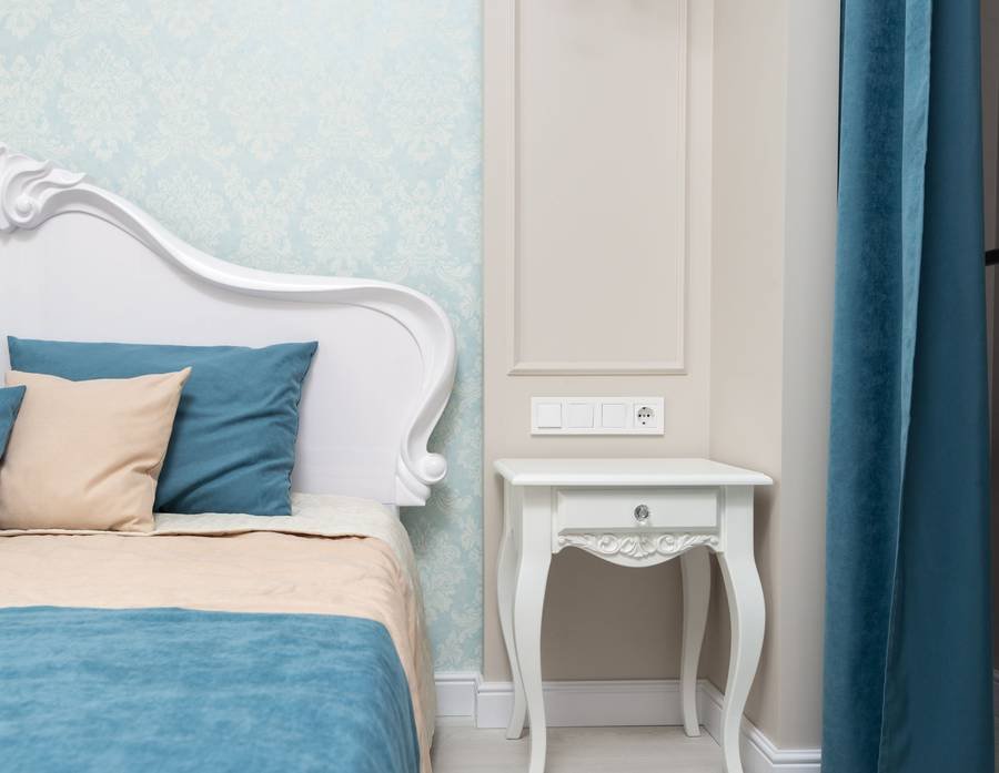 A white French-style bed against a moulded wall, with teal bedding and a curved nightstand