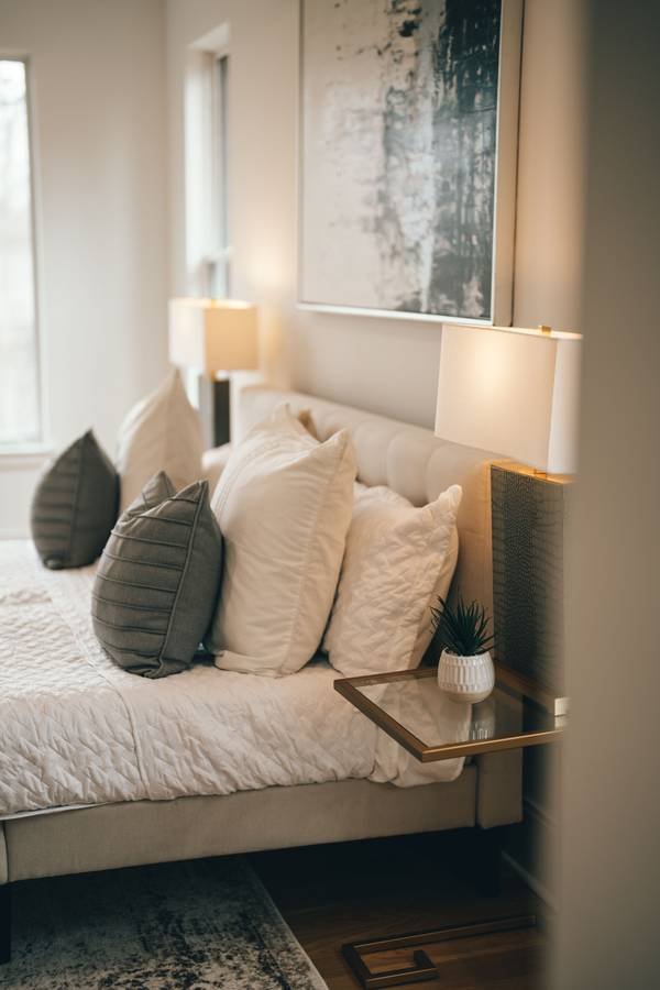 Layered cushions on a bed lit by a bedside lamp, with a large abstract painting above